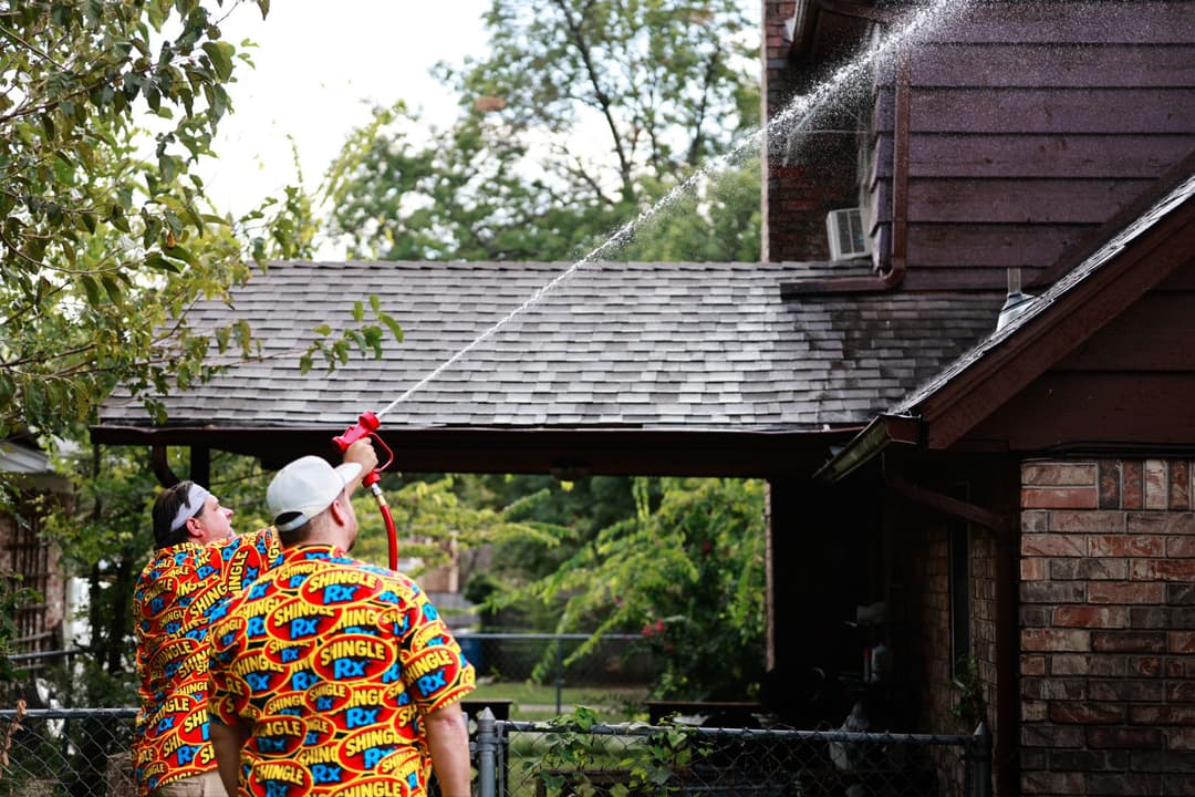 Two individuals in colorful shirts spraying water on a house roof with a hose.
